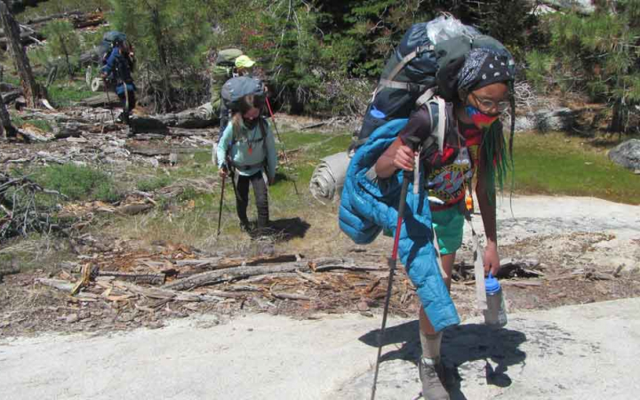 Students wearing backpacks make their way across a grassy and rock landscape. They brace themselves with hiking poles.